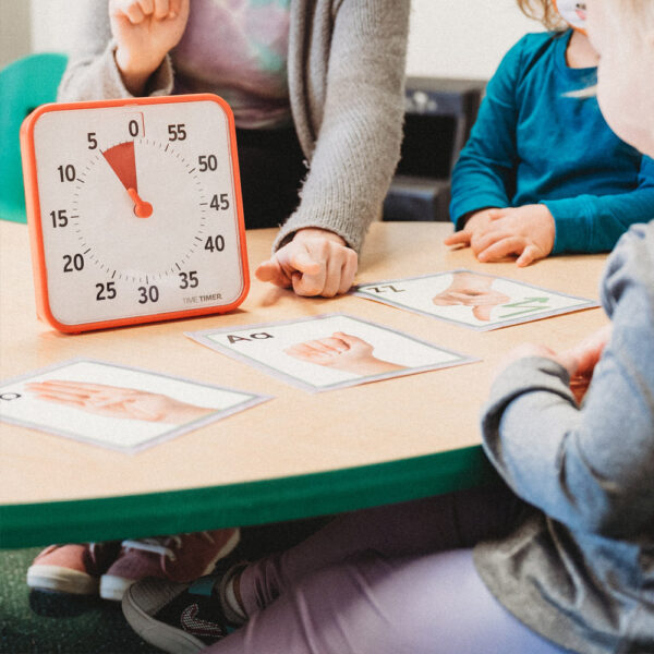 En vuxen pekar på ett bildkort som ligger bredvid en orange Time Timer Medium och barn tittar på